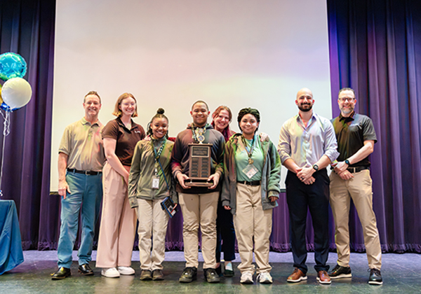 A group of students and adults pose on a stage holding a trophy plaque at an awards ceremony, with blue and gold balloons visible in the background.