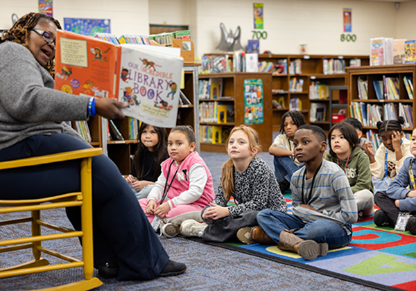 A teacher reads aloud Our Incredible Library Book to a group of diverse elementary students sitting on a colorful rug in a school library.